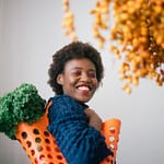 happy young black woman holding basket with lettuce on shoulder and cluster of yellow dates in grocery store on gray background
