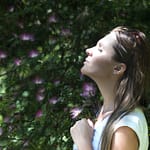 woman closing her eyes against sun light standing near purple petaled flower plant