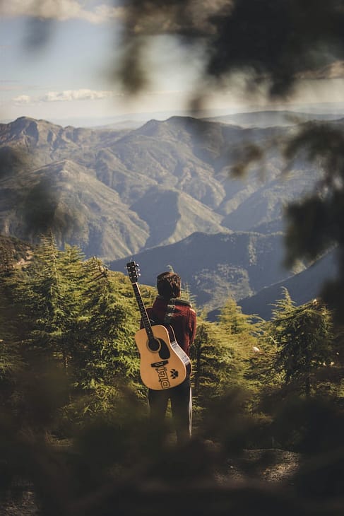 person with acoustic guitar standing in green field near mountain